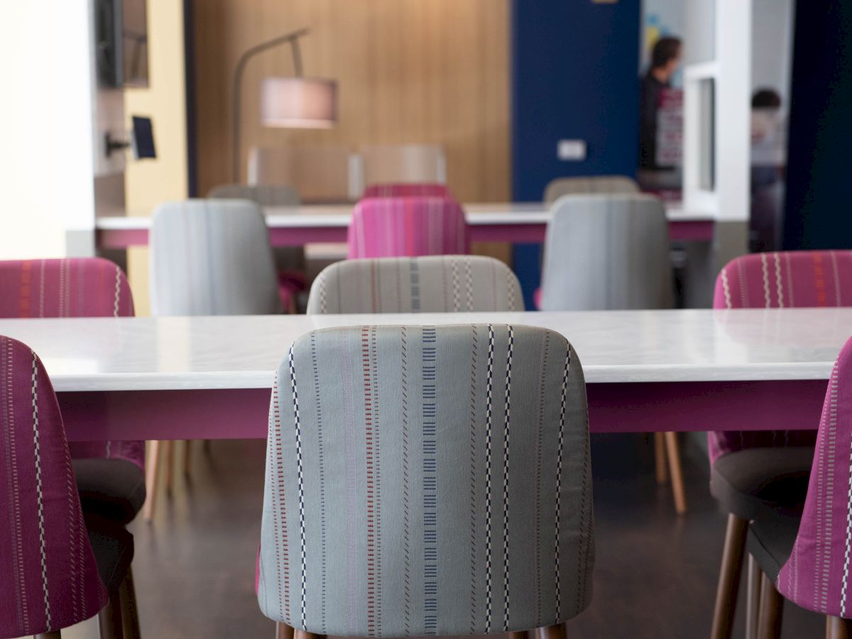 The image shows a modern dining area with a row of colorful chairs around white tables. The lighting is warm, and the decor is contemporary.