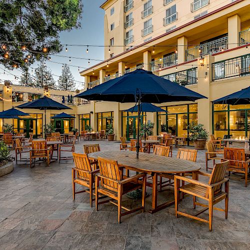 An outdoor dining area with wooden tables and chairs, blue umbrellas, string lights, and a yellow building in the background.