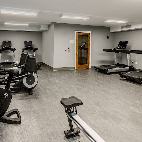 A gym with various equipment, including treadmills, stationary bikes, and a rowing machine, all in a well-lit room with gray walls and flooring.