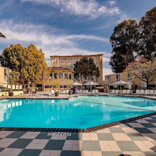 A large outdoor swimming pool surrounded by lounge chairs and umbrellas, with a building in the background and trees under a partly cloudy sky.