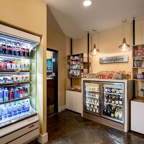 The image depicts a small convenience store area with refrigerated beverages, snacks, and an assortment of other items on shelves, along with a checkout counter.