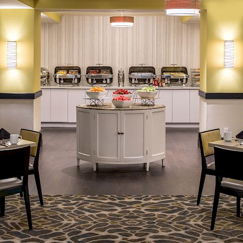 The image shows a dining area with neatly set tables, a central island holding fruit bowls, and a buffet with covered trays in the background.