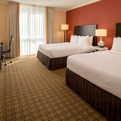 The image shows a tidy hotel room with two double beds, lamps, a TV, a desk with a chair, and a large window with curtains allowing natural light to enter.