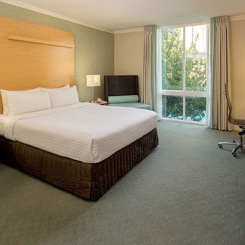 This image shows a hotel room with a double bed, side tables, lamps, TV, desk, chair, and large window with curtains.