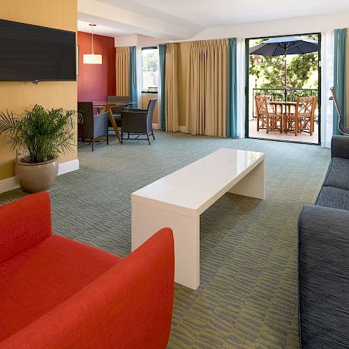 Spacious living room with modern furniture, including a red chair, blue sofa, white table, mounted TV, and patio doors leading to an outdoor dining area.