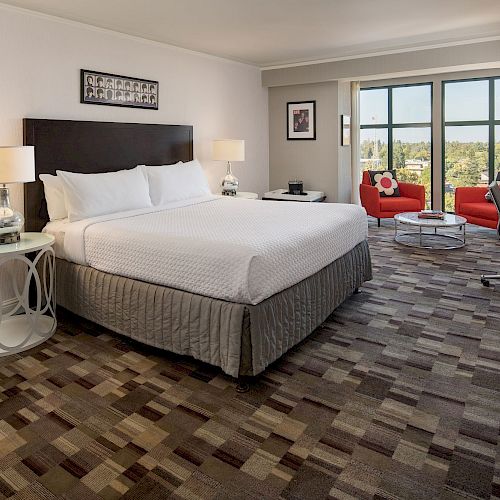 This image shows a modern hotel room with a large bed, lamps, red chairs by the window, a TV, and a turntable. The room has a neutral color scheme.