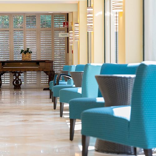 The image shows a bright hallway with blue chairs lined up along glass windows on the right, and a wooden table with a plant near the end.