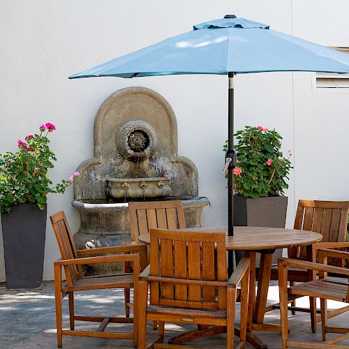 An outdoor patio with wooden furniture, a round table, chairs, a blue umbrella, potted plants, and a wall-mounted decorative water fountain.