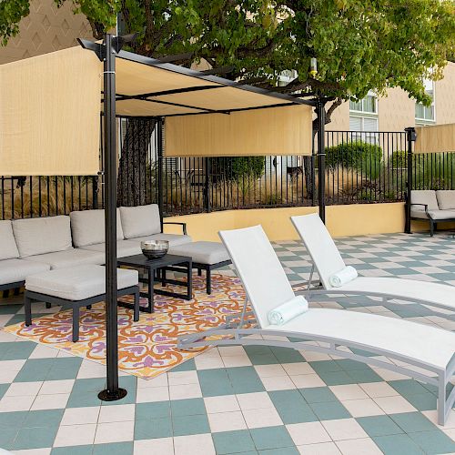 An outdoor lounging area with cushioned seats, chaise lounges, and canopy shelters on a checkered tile floor, near a building with surrounding greenery.