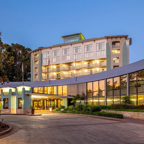 The image shows the exterior of a modern hotel with a curved entrance, large windows, and a lit facade, set against a clear blue sky.