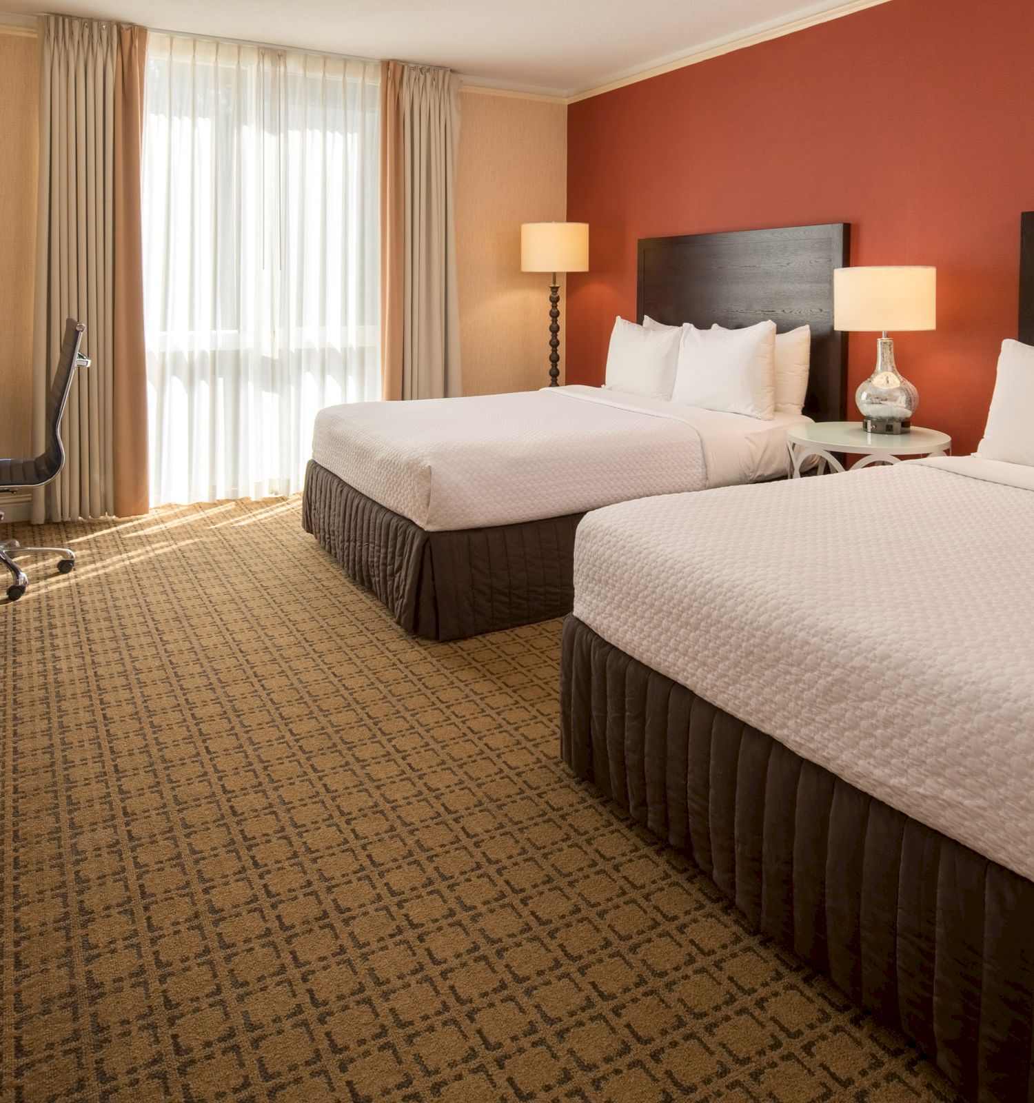 The image shows a hotel room with two queen beds, bedside tables, lamps, a desk, chair, TV on a cabinet, and large window with curtains.