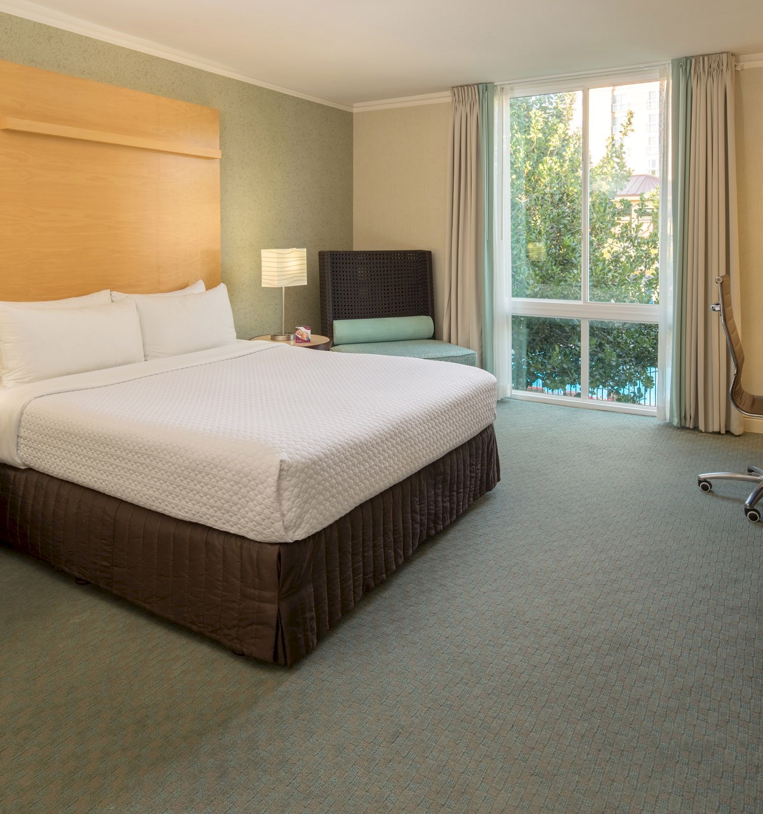 A hotel room with a king-sized bed, side tables with lamps, a TV, desk, chair, sofa by the window, and greenery visible outside.