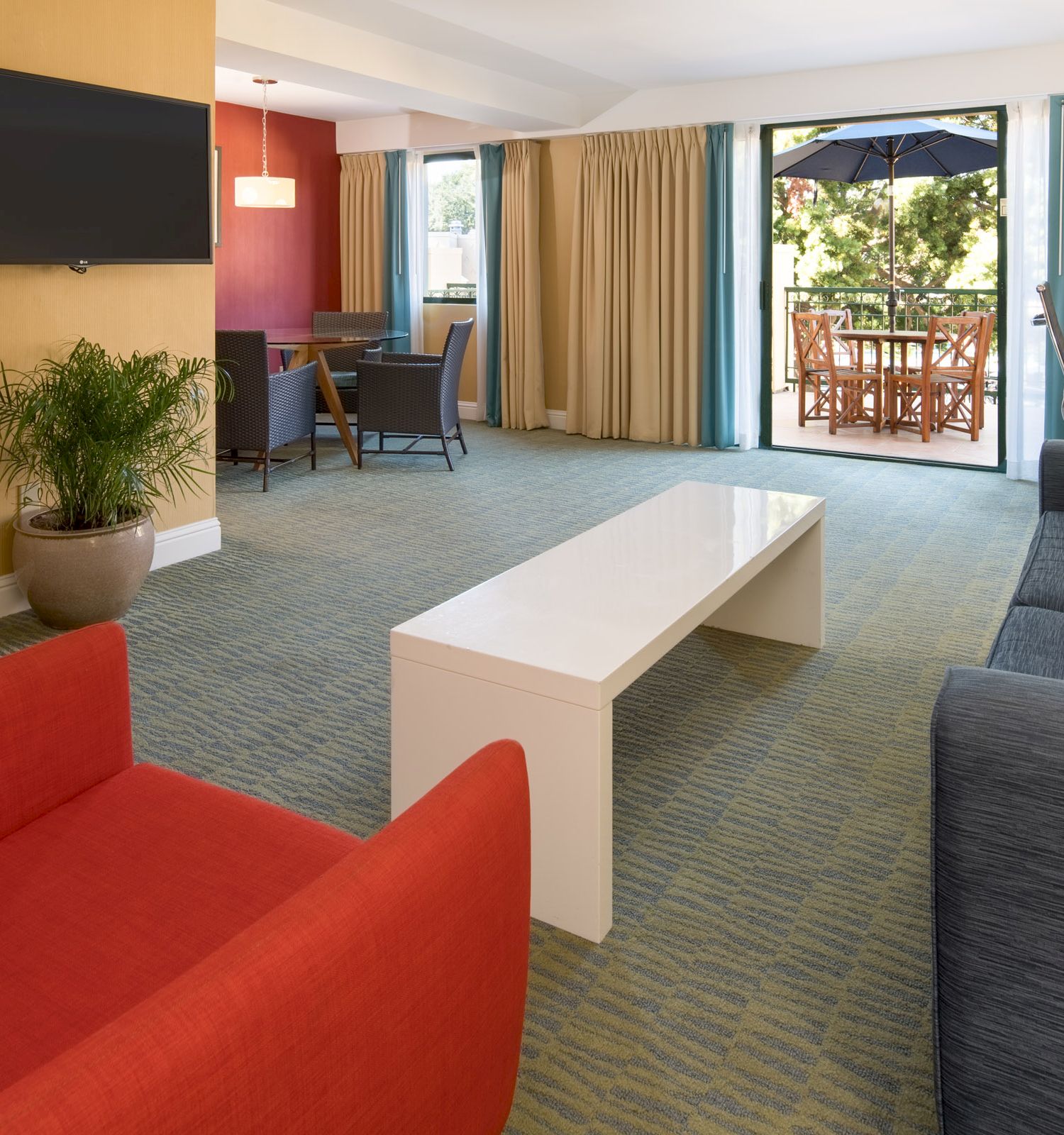 The image shows a modern living room with a red chair, grey sofa, white coffee table, wall-mounted TV, and a dining table, opening to a patio area.