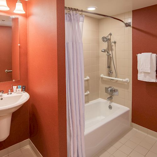 A bathroom with a pedestal sink, mirror, toiletries, bathtub with shower, white curtain, and towel rack with towels. Ends the sentence.