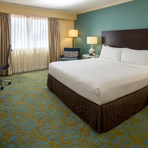 A hotel room featuring a king-sized bed, nightstands with lamps, a TV, a desk with a chair, and a window with curtains.