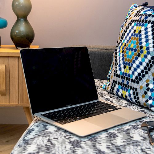 A laptop on a patterned bedspread, with a patterned pillow, next to a wooden side table with books, a camera, and two lamps.