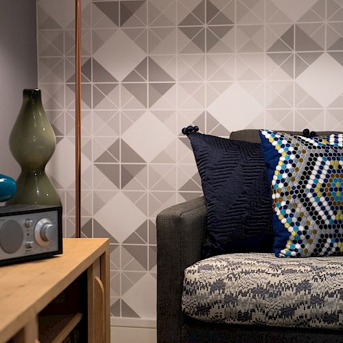 A cozy living room corner with a patterned couch, geometric pillows, a wooden table holding vases, and a radio, against a geometric wallpaper.