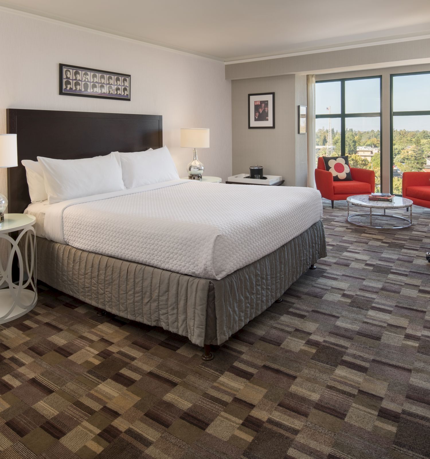 This image shows a hotel room with a large bed, two nightstands with lamps, a TV, a seating area with red chairs, and a window with a scenic view.