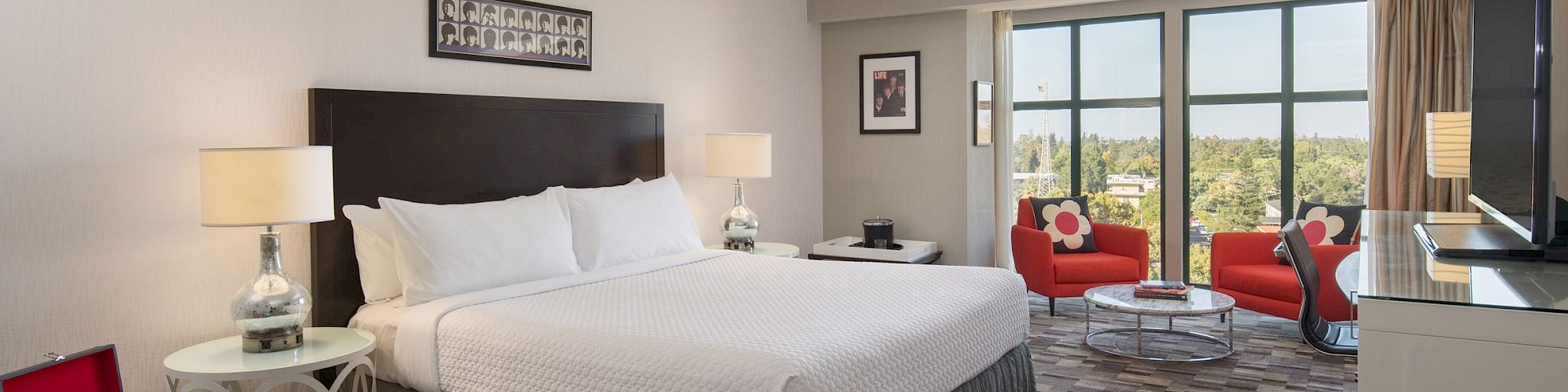 The image shows a modern hotel room with a large bed, bedside tables, a TV, and a seating area with two red chairs near the window.