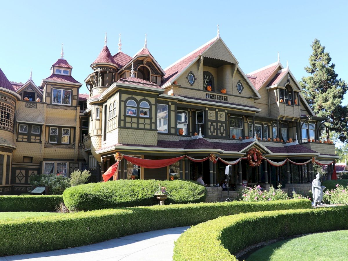 A large, ornate Victorian-style mansion with multiple gables and decorative windows surrounded by well-manicured gardens and pathways.