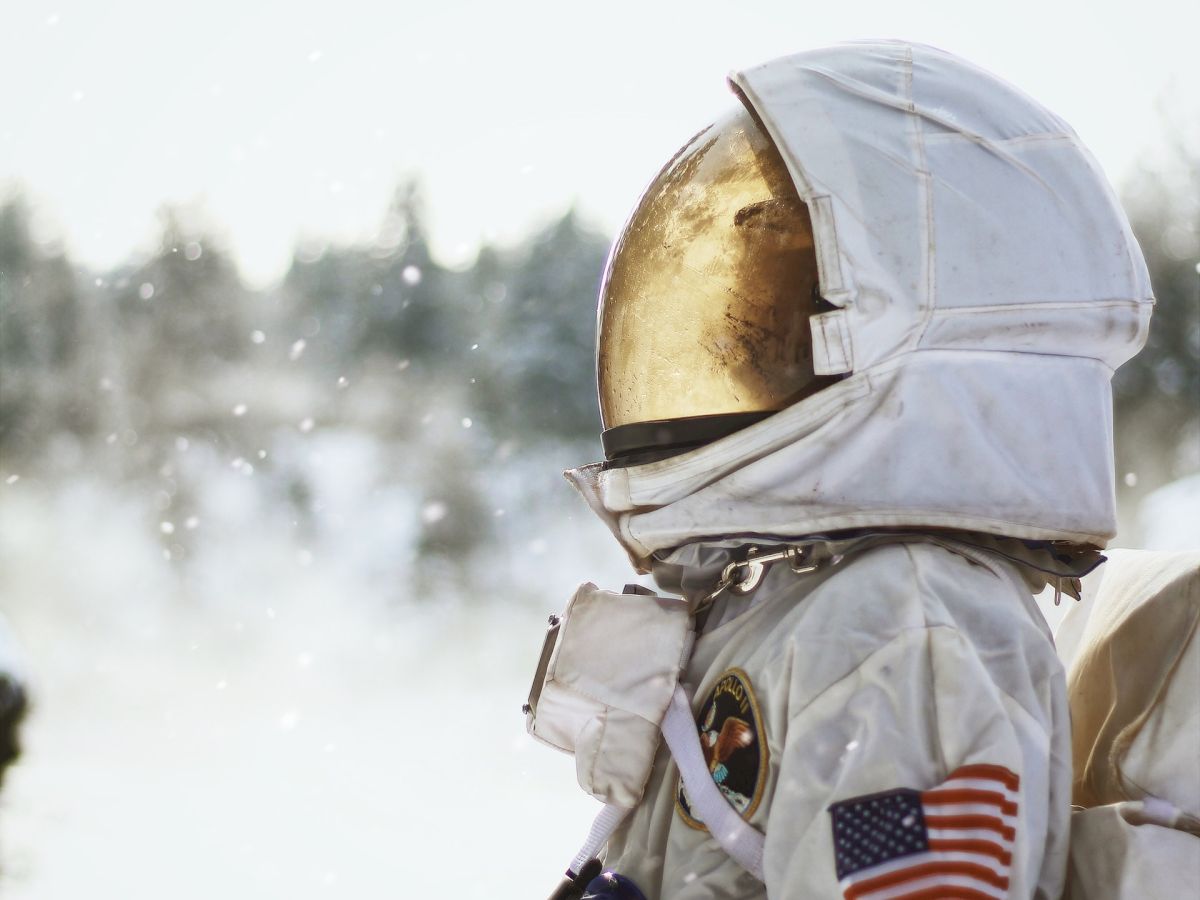 A person in a spacesuit with a reflective helmet stands outdoors in a snowy environment, with trees and snow visible in the background.