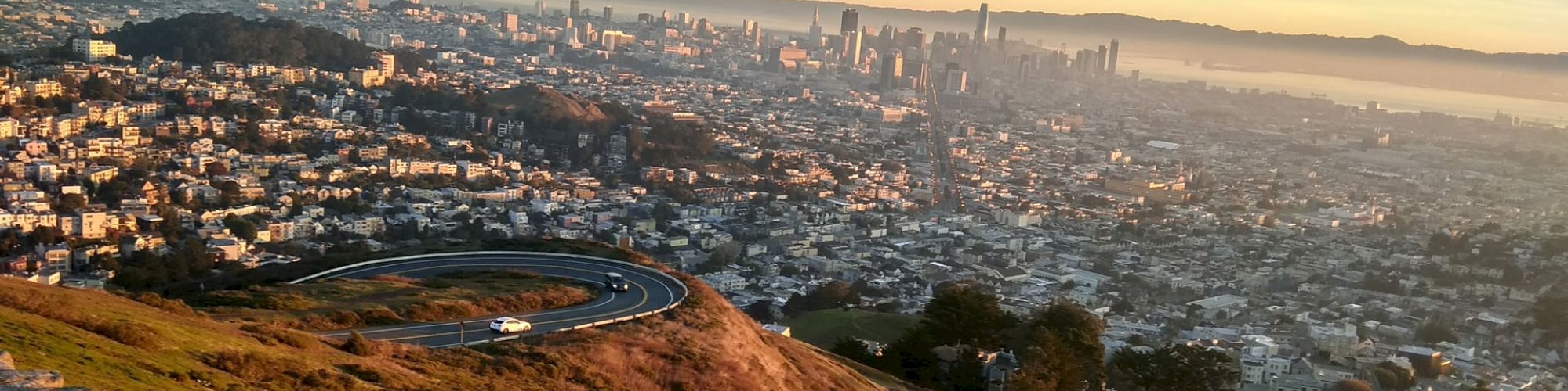 A scenic view of a city with numerous buildings, taken from a high vantage point at sunset, showing a winding road and a car on a hillside.