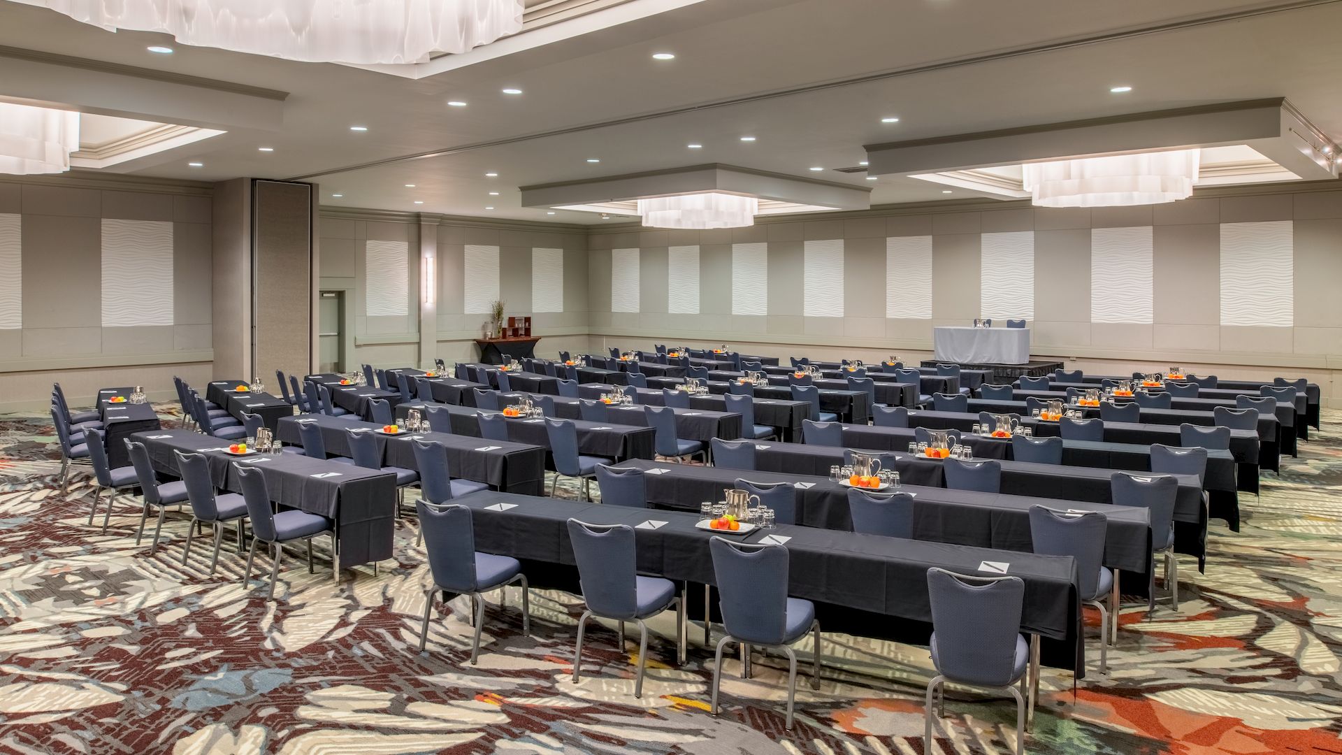 The image shows a conference room set up with multiple rows of tables and chairs, facing a stage area with a podium and equipment.