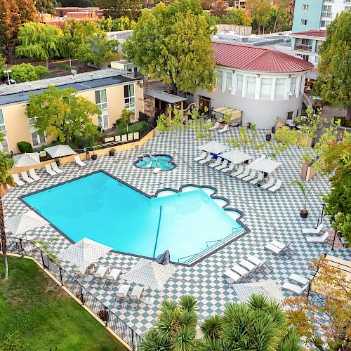 An aerial view of a large, uniquely-shaped swimming pool surrounded by lounge chairs and umbrellas. Nearby is a hot tub, trees, and buildings.