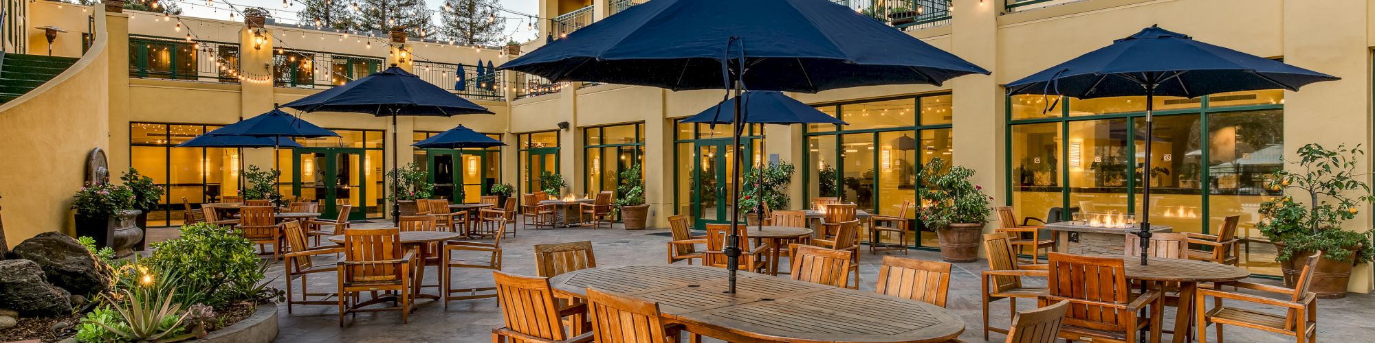 The image shows an outdoor patio area with wooden tables and chairs, shaded by blue umbrellas, and surrounded by a multi-story building with balconies.