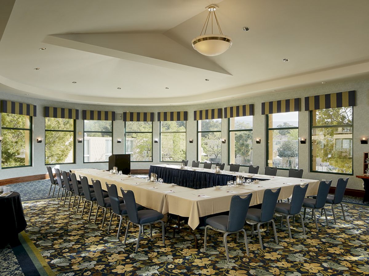 The image shows an empty conference room with a U-shaped table setup, surrounded by windows, adorned with striped curtains, under a large ceiling light.