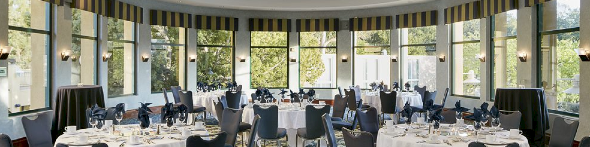 The image shows a dining room with round tables covered in white linens, set with dishware and surrounded by gray chairs, under a ceiling light.