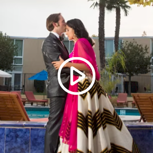 A couple in elegant attire is embracing near a pool with deck chairs and trees in the background. The woman wears a colorful traditional dress.