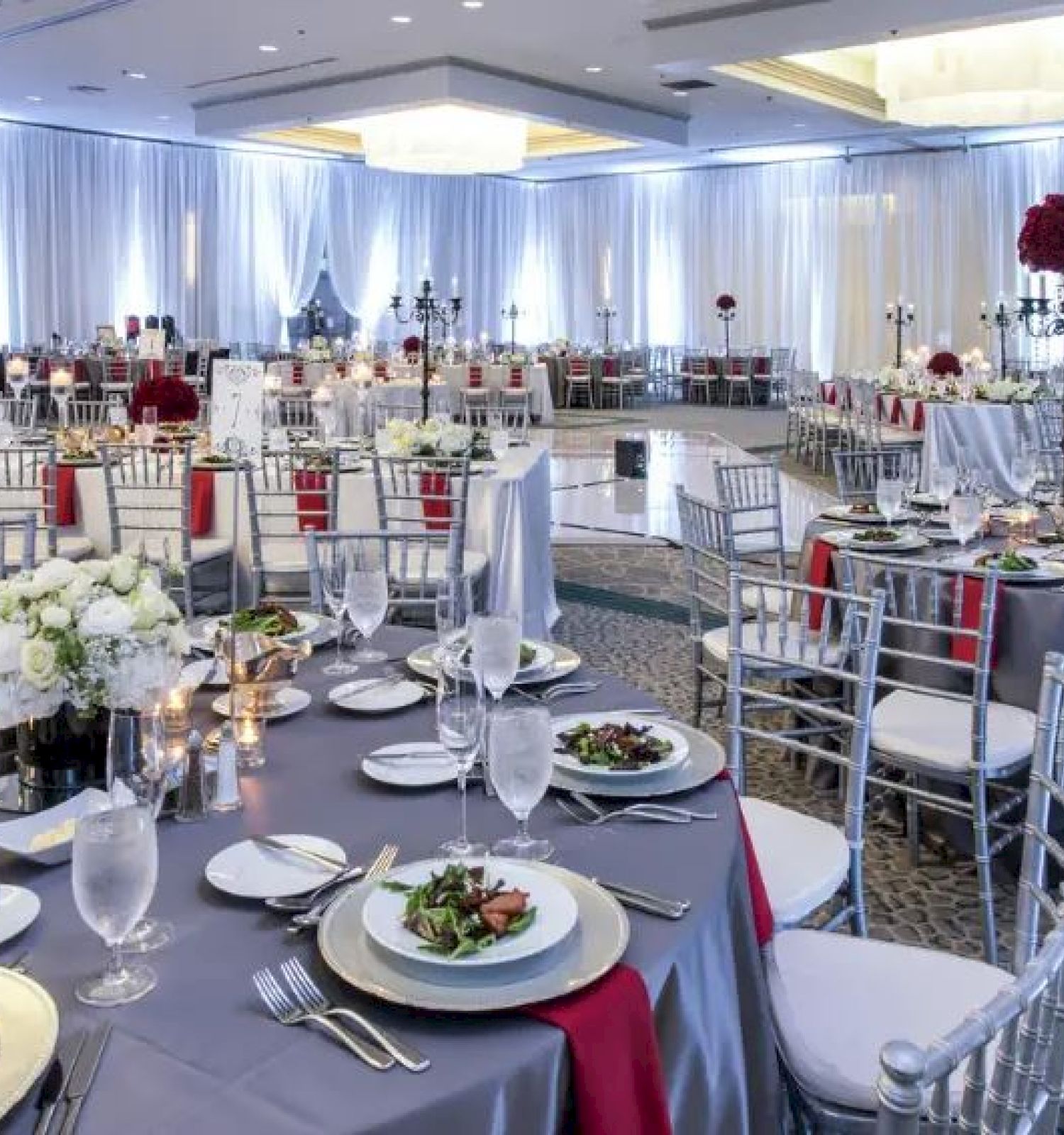 The image shows a luxurious, decorated indoor banquet hall set up for an event, with round tables arranged with plates, utensils, and flower centerpieces.