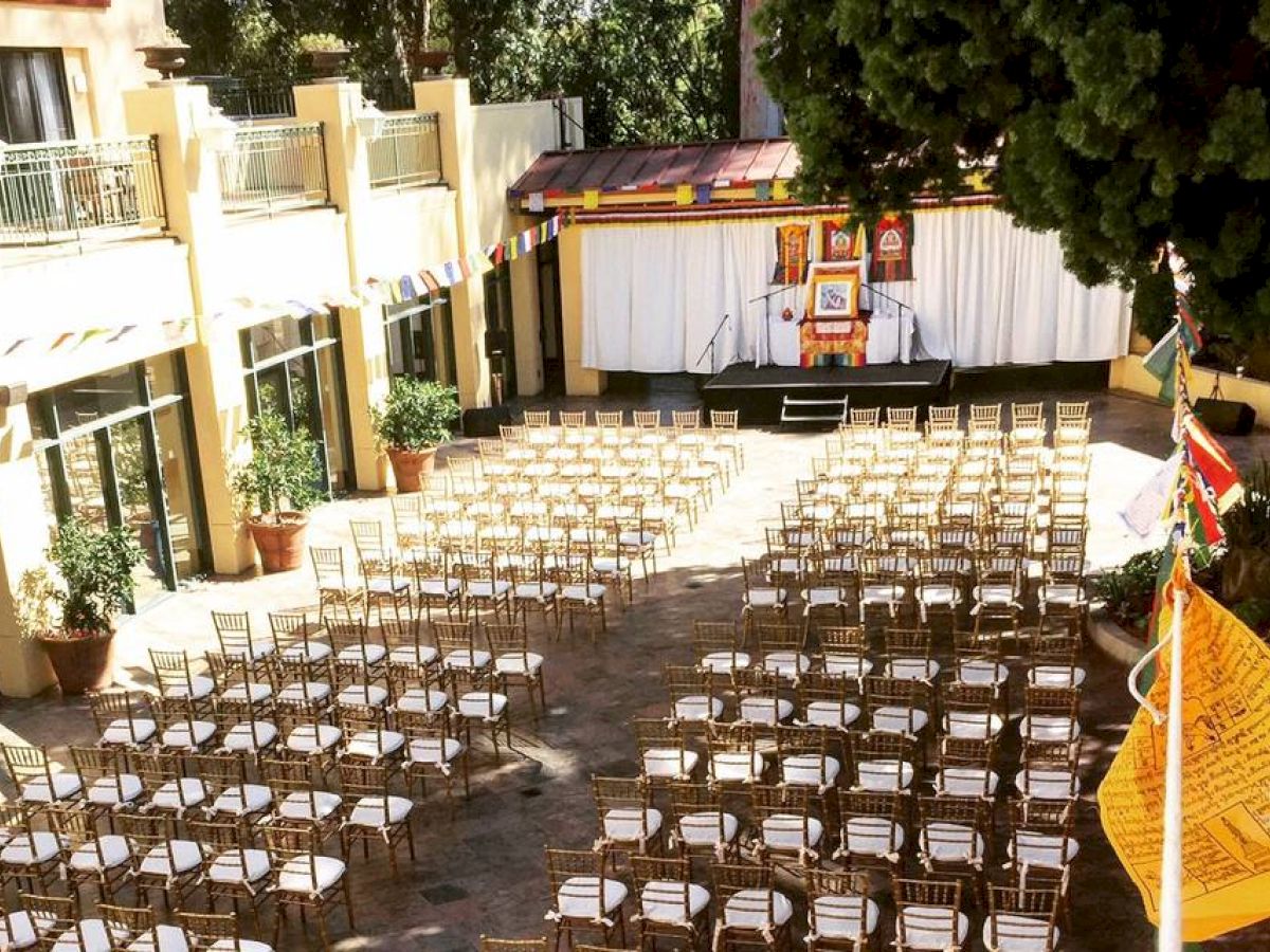 An outdoor event setup with rows of chairs facing a stage, surrounded by buildings and greenery, under a sunny sky with colorful flags hanging.