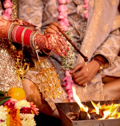 A wedding ceremony where a couple is performing rituals near a fire, with the bride's henna-decorated hands and jewelry visible.