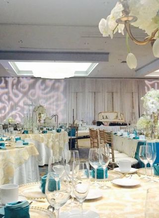 The image shows an elegant banquet hall with tables set up for an event, decorated with white flowers, blue napkins, and gold accents, under soft lighting.