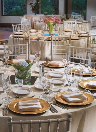 Elegantly set tables with white cloths and gold-rimmed plates, ready for a formal event, with floral centerpieces and neatly arranged cutlery.