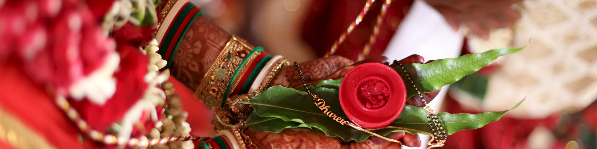 The image shows a person's hands adorned with henna and bangles, holding a red rose and green leaves, likely during a traditional ceremony.