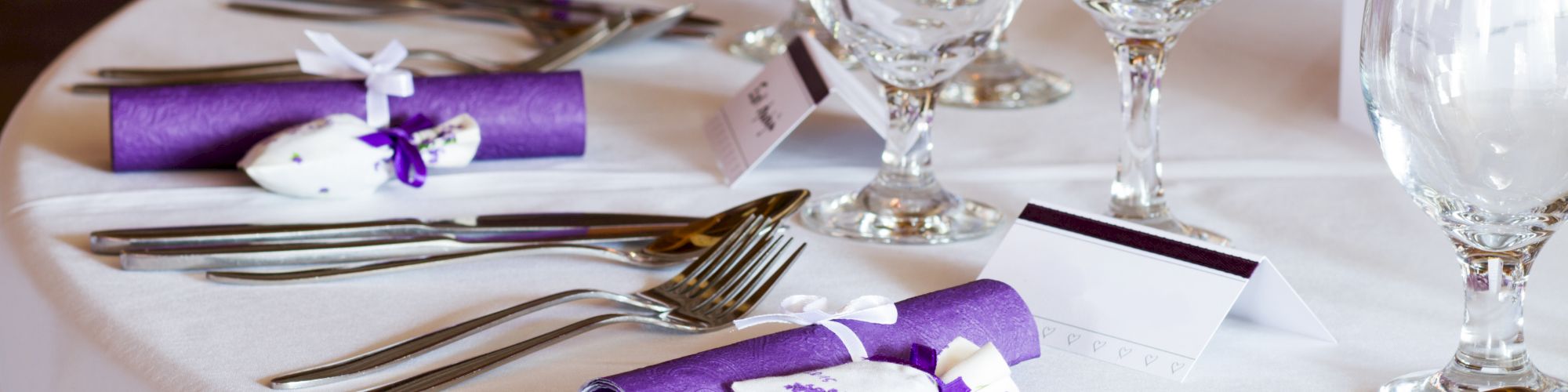 A elegantly set table with purple napkins, name cards, wine glasses, and utensils arranged neatly on a white tablecloth.