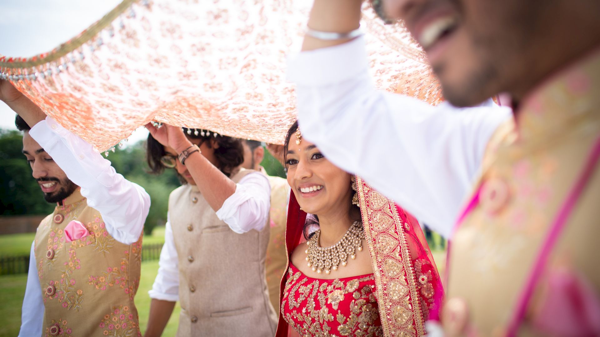 A bride walks under a decorated canopy, smiling, accompanied by three people in traditional attire, during an outdoor celebration or ceremony.