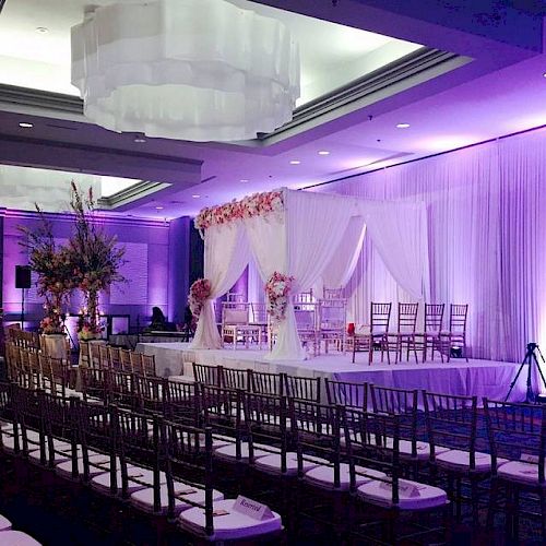 A beautifully decorated wedding venue features a draped ceremony backdrop, flowers, rows of chairs, and purple lighting, creating an elegant atmosphere.