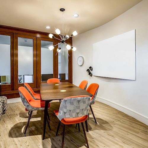 A modern conference room with a wooden table, orange chairs, wall art, a whiteboard, and a unique light fixture, including a refreshment area.