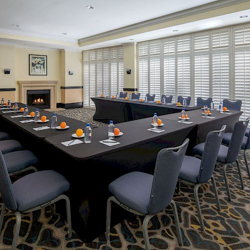 A conference room with a U-shaped table setup, chairs, water bottles, pens, notepads, and oranges, complemented by a fireplace and large windows.