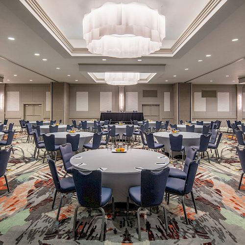 A spacious, elegantly lit banquet hall with round tables and chairs set up, ready for an event or conference, featuring modern decor and chandeliers.