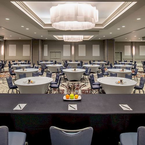 A conference room setup with round tables, blue chairs, and a stage with note cards and fruit. The room has modern lighting and neutral decor.