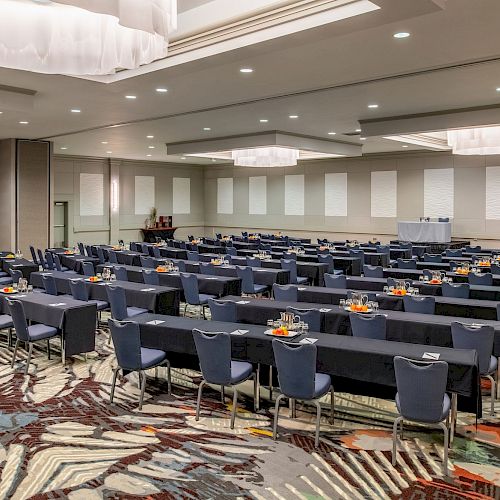 A conference room is set up with rows of tables and chairs, each with notepads and water. The room features modern decor, carpeting, and ceiling lights.