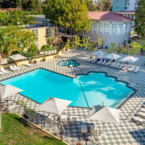 This image shows an outdoor swimming pool area with a unique shape, surrounded by lounge chairs, umbrellas, and palm trees.