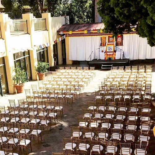 This image shows an outdoor event setup with many rows of chairs facing a stage with a backdrop, surrounded by buildings and greenery, ending the sentence.