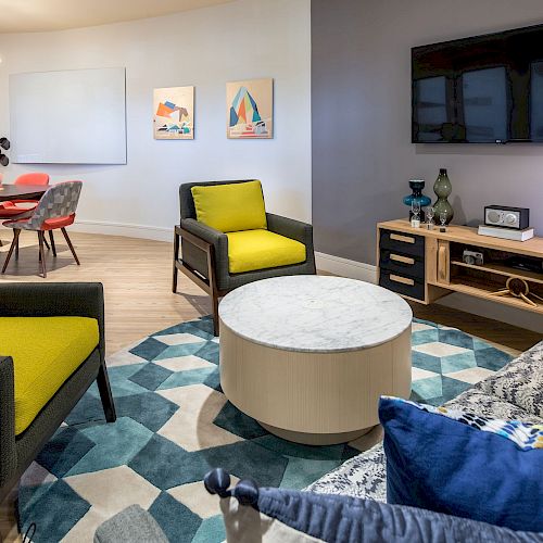 A modern living room with a TV, armchairs, a sofa, round table, and a geometric-patterned rug; dining table and chairs are visible in the background.