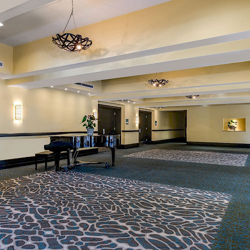 This image shows a spacious, well-lit hotel hallway with patterned carpets, chandeliers, a piano, and decor along the walls.